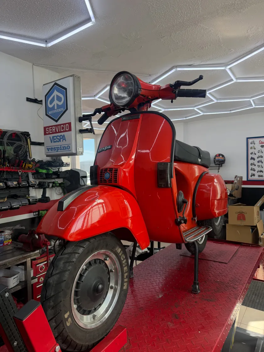 La misma moto acabada en rojo brillante, sobre el elevador y bajo las luces hexagonales del taller.