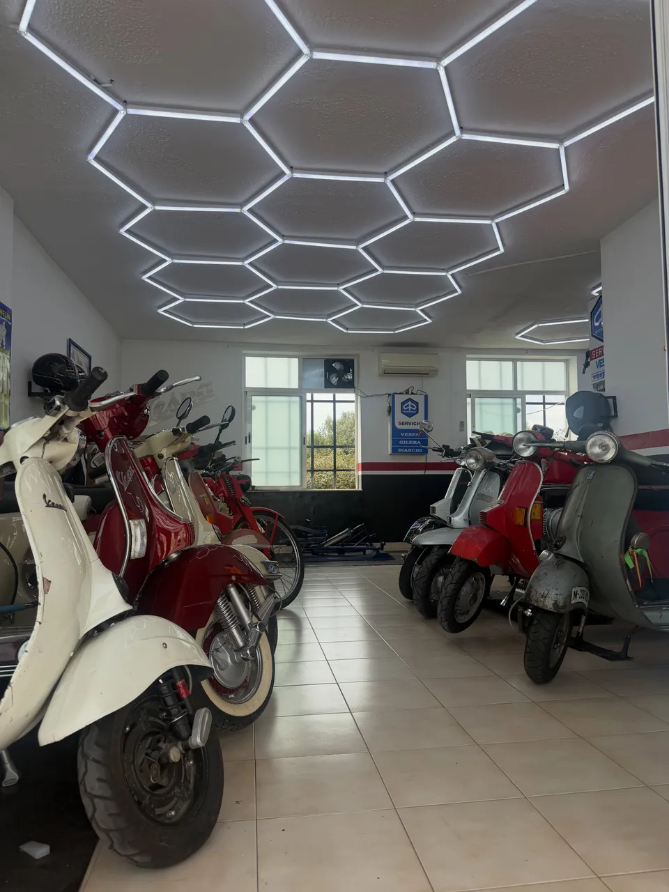 Interior del taller, con Vespas clásicas aparcadas a ambos lados y los paneles LED hexagonales encendidos en el techo.
